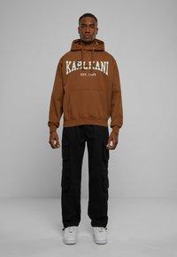 Felpa con cappuccio marrone, con testo bianco "KARL KANI EST. 1989" e tasca frontale, abbinata a pantaloni cargo neri e sneakers bianche.
