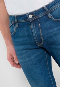 Jean en denim bleu foncé à coupe slim, avec des coutures blanches, une fermeture à bouton à l'avant, et des poches latérales inclinées.