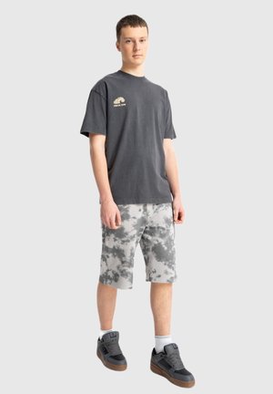 Młody mężczyzna stoi, ubrany w czarny T-shirt z nadrukiem, szaro-białe spodnie szorty tie-dye, białe skarpetki i czarne trampki na jednolitym tle.