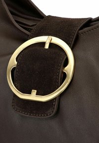 Borsa in pelle marrone con una tracolla in scamosciato marrone scuro e una grande fibbia circolare dorata che valorizza la chiusura. Materiali testurizzati visibili.