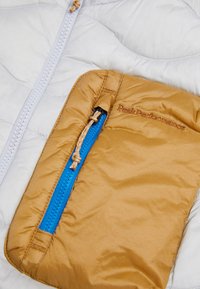 Lätt, vit quiltad jacka med en guldplånbok som har en blå dragkedja. "Peak Performance"-logotypen präglad på fickan.