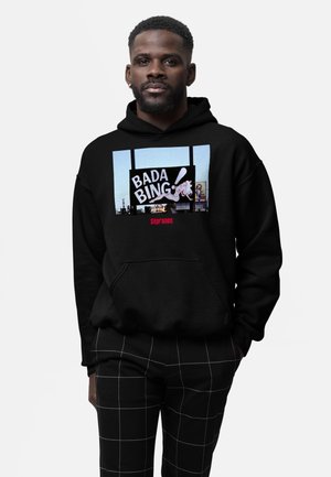 Man met zwarte hoodie met "Bada Bing!" bordgrafiek en "Sopranos" tekst, gecombineerd met zwarte broek met witte rasterlijnen.