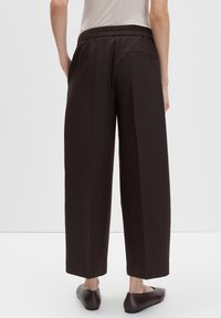 Personne portant un pantalon ample marron foncé avec une taille élastique et des chaussures plates noires, se tenant debout contre un fond clair uni.