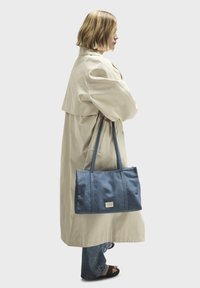 Femme blonde en trench-coat beige et jeans, debout de profil, tenant un grand sac fourre-tout en denim bleu avec de longues anses.
