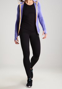Felpa zip a viola con accentuazioni arancioni, canotta nera aderente, leggings neri e scarpe sportive nere con dettagli turchesi.