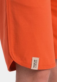 Shorts en coton orange avec une texture lisse, présentant des ourlets cousus et une étiquette en bas de l'ourlet affichant le texte "VOTRE SEUL LIMIT EST VOTRE ESPRIT."