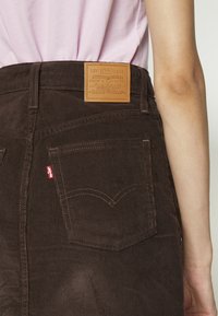 Vue de dos d'une personne portant une jupe en velours côtelé marron avec un patch en cuir Levi's et une étiquette rouge, associée à un haut rose clair.