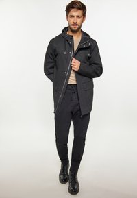 Chaqueta impermeable negra con cremallera frontal, capucha ajustable y grandes bolsillos frontales. Lleva pantalones casuales negros y botas negras con cordones.
