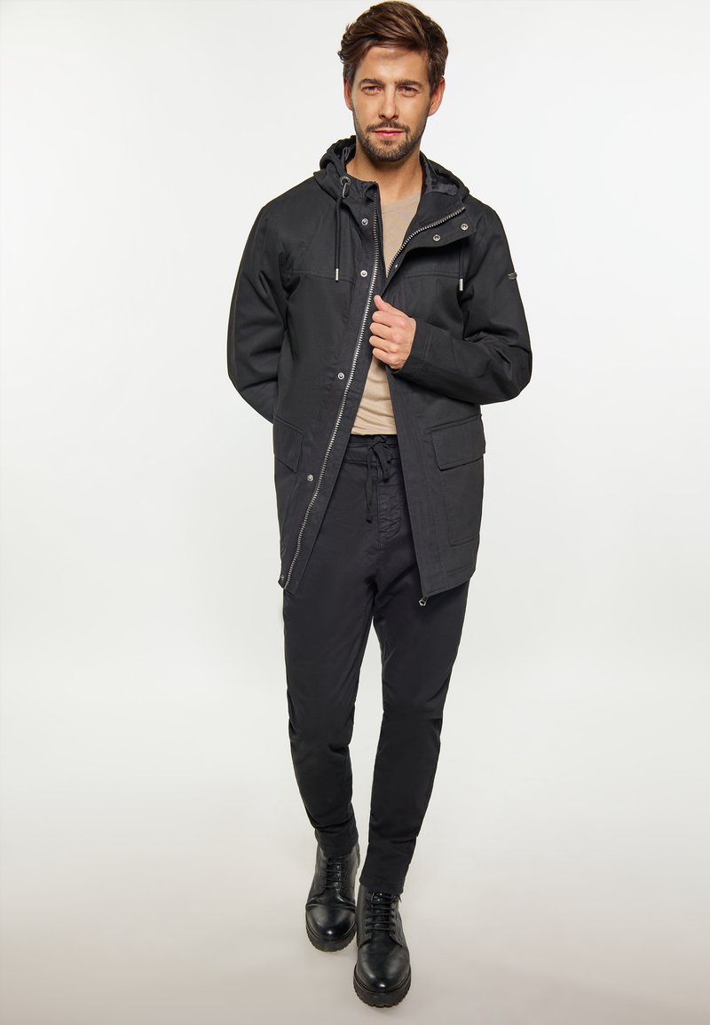 Schwarze wasserdichte Jacke mit durchgehendem Reißverschluss, verstellbarem Kapuze und großen Fronttaschen. Getragen mit schwarzen Freizeitpants und schwarzen Schnürstiefeln.