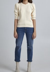 Cremfarbener, gestrickter Pullover mit voluminösen kurzärmeligen Ärmeln, runder Ausschnitt und geripptem Saum. Getragen mit ausgebleichten blauen Jeans und beigen Knöchelstiefeln.