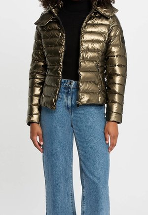 Femme portant une doudoune bronze métallique sur un col roulé noir et un jean taille haute bleu clair à jambes larges, debout devant un fond uni.