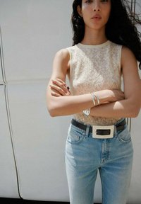 Lace tanktop i creme, lyse blå jeans med høj talje, sort bælte med sølvspænde og sølvarmbånd, arme korslagte.