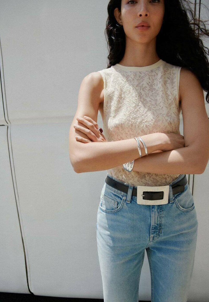 Lace tanktop i creme, lyse blå jeans med høj talje, sort bælte med sølvspænde og sølvarmbånd, arme korslagte.