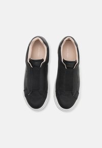 Zapatos negros de cuero tipo slip-on con paneles elásticos, suelas de goma blancas y un forro beige. Presentan una marca mínima en la parte superior.