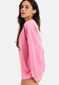 Rosa oversized hettegenser med teksturert veving, med lange ermer og grafisk trykk på fronten.