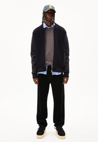 Blouson de pilote navy sur un pull marron, chemise rayée bleu clair, pantalon en velours côtelé noir, chaussures noires avec semelles beiges, casquette grise avec logo jaune.