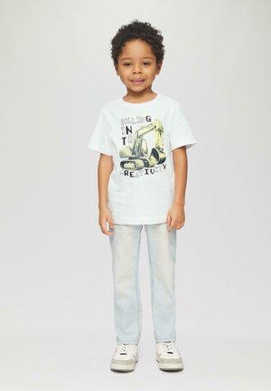 Witte katoenen t-shirt met een graafmachine grafiek en tekst, gedragen met lichtblauwe denim jeans en witte schoenen met grijze accenten.