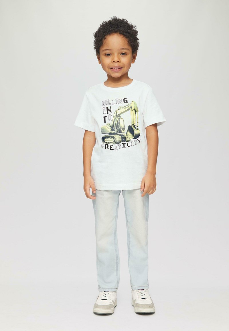 Weißes Baumwoll-T-Shirt mit einem Bagger-Graphic und Text, kombiniert mit hellblauen Jeans und weißen Schuhen mit grauen Akzenten.
