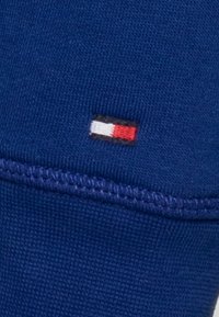 Tejido azul con una textura acanalada y un pequeño logo bordado en negro, blanco y rojo en la costura.