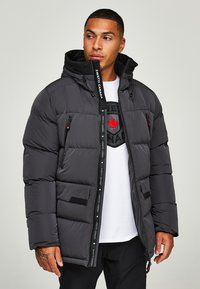 Donkergrijze gewatteerde jas met capuchon, voorzien van zipzakken, een zwarte ritssluiting en een rood esdoornbladlogo op de borst.