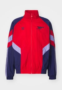 Crvena i mornarsko plava jakna s visokim ovratnikom, zatvaranjem na zatvarač i elastičnim manžetama. Ima bijele i svijetlopljive pruge i logotipe.