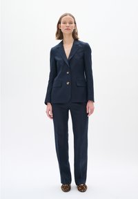 Námornícky lněný blazer a odpovídající kalhoty s hladkou texturou. Blazer má dva velké knoflíky a kapsy. Osoba má na sobě boty s potiskem zvířat.