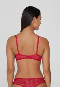 Červená krajková podprsenka s nastavitelnými ramínky a háčkovým zapínáním na zádech, zdobená květinovými vzory, spolu s odpovídajícími krajkovými kalhotkami ve stejné barvě.