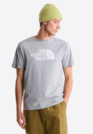 Camiseta de algodón gris con un gran logo blanco en el pecho, combinada con pantalones kaki y un gorro de punto texturizado de color verde claro.