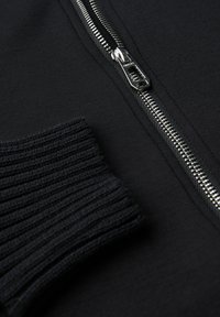 Giacca nera con zip e polsini a coste, dotata di zip metallica e superficie in tessuto liscio. La prospettiva ravvicinata mette in evidenza i dettagli.