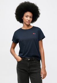 Γυναίκα με αφρο χτένισμα, φορώντας μπλε μπλούζα με μικρή κόκκινη καρδιά και μαύρα τζιν, χέρι στη τσέπη, ουδέτερο φόντο.