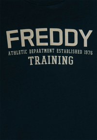 Tessuto scuro con testo in grassetto "FREDDY," testo più piccolo "DEPARTAMENTO ATLETICO FONDATO NEL 1976," e "ALLENAMENTO" sottostante in lettere bianche.