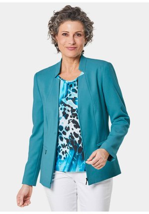 Teejacket in Teal mit strukturiertem Design, langen Ärmeln, vorderen Taschen und gerader Silhouette, kombiniert mit einem blauen gemusterten Oberteil und weißen Hosen.