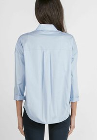 Hellblaues Button-up-Hemd mit einem Rückenfalten, hochgekrempelten Ärmeln und einem abgerundeten Saum. Hergestellt aus einem glatten Stoff. Einfaches Kragen-Design.