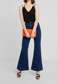 Top camicia nero con spalline sottili, jeans svasati blu scuro a vita alta e pochette arancione. La modella indossa sandali neri con cinturini.