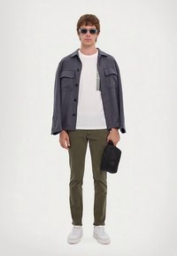 Moška obleka zivo sivo srajco s gumbi, belo majico s kratkimi rokavi in žepom, olivno zelenimi hlačami in belimi supergami, v roki drži črno kapo.