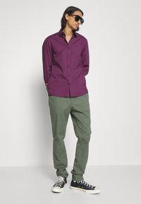 Camicia viola a maniche lunghe con chiusura a bottoni, abbinata a pantaloni verde oliva e sneakers nere. Occhiali da sole indossati, mani in tasca.