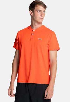 Giovane uomo che indossa una maglietta arancione brillante a maniche corte con due bottoni e il logo bianco "nox", abbinata a pantaloncini neri, che guarda verso destra.