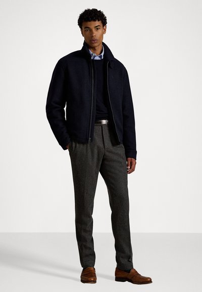 Giacca a zip blu marina con colletto alto, indossata sopra un maglione scuro e una camicia a fantasia, abbinata a pantaloni grigi e mocassini marroni.