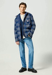 Chaqueta de cuadros azules sobre un chaleco marino, combinada con jeans de color azul claro y zapatos negros. Presenta un gran patrón de cuadros y bolsillos frontales.