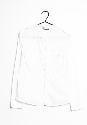 Camisa blanca de manga larga con cierre de botones al frente, tapeta con volantes, dos bolsillos en el pecho y charreteras en los hombros. Textura suave.