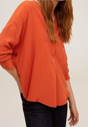 Femme portant un chemisier orange ample à manches longues et un jean foncé, debout avec une main dans le dos.