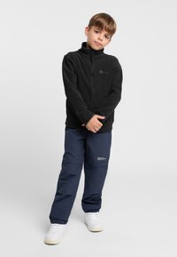 Veste en polaire noire avec col montant, fermeture éclair à l'avant et logo. Associée à un pantalon bleu marine et des baskets blanches avec semelles en caoutchouc.