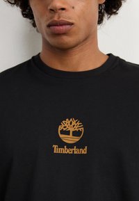 Schwarzes Baumwoll-T-Shirt mit einem goldenen Timberland-Logo, das ein Baumdesign und darunter stehenden Text zeigt, runder Halsausschnitt und kurze Ärmel.