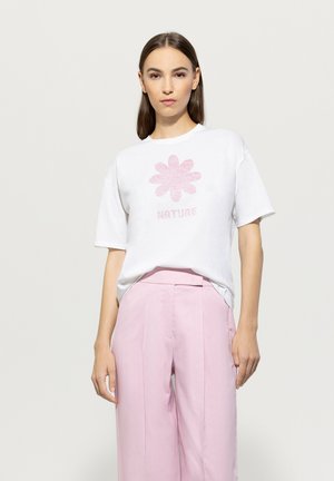 Weißes T-Shirt mit kurzen Ärmeln, das ein pinkes Blumenmotiv und das Wort "NATURE" zeigt. Kombiniert mit hellrosa hochgeschnittenen Hosen.