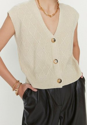 Beige gebreid mouwloos vest met drie knopen, gedragen met een zwarte broek, gouden ketting en armbanden.
