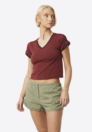 Młoda kobieta o krótkich blond włosach, ubrana w brązowy krótki T-shirt z dekoltem w serek oraz oliwkowozielone szorty z elastycznym pasem, patrząca na bok.