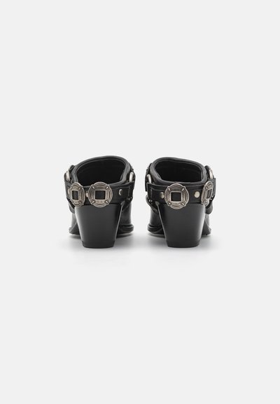 Zuecos de tacón de cuero negro con punta redonda, correa trasera y acentos de hardware plateado con diseños circulares en las hebillas.