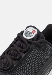 Crne atletske tenisice s mrežastim gornjim dijelom, teksturiranim uzorkom i ravnim vezicama. Imaju okrugli logo na jeziku s natpisom "AIR MAX".