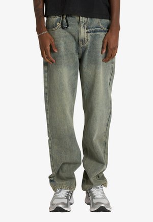 Persona che indossa jeans blu sbiaditi e a vestibilità ampia, braccialetto d'argento, top nero e scarpe da ginnastica grigie e bianche, in piedi con le mani rilassate ai lati.