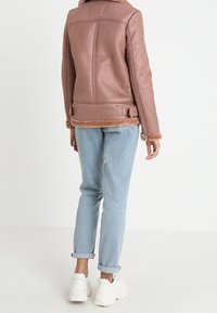Giacca di pelle rosa con rifiniture in shearling, caratterizzata da un taglio dritto e una cintura regolabile. Indossata con jeans azzurri chiari e sneakers bianche.